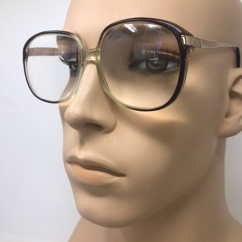 May include: Vintage eyeglasses with a brown and clear frame. The glasses have a square shape and a gold-colored metal arm. The lenses are clear and reflect the surrounding environment.