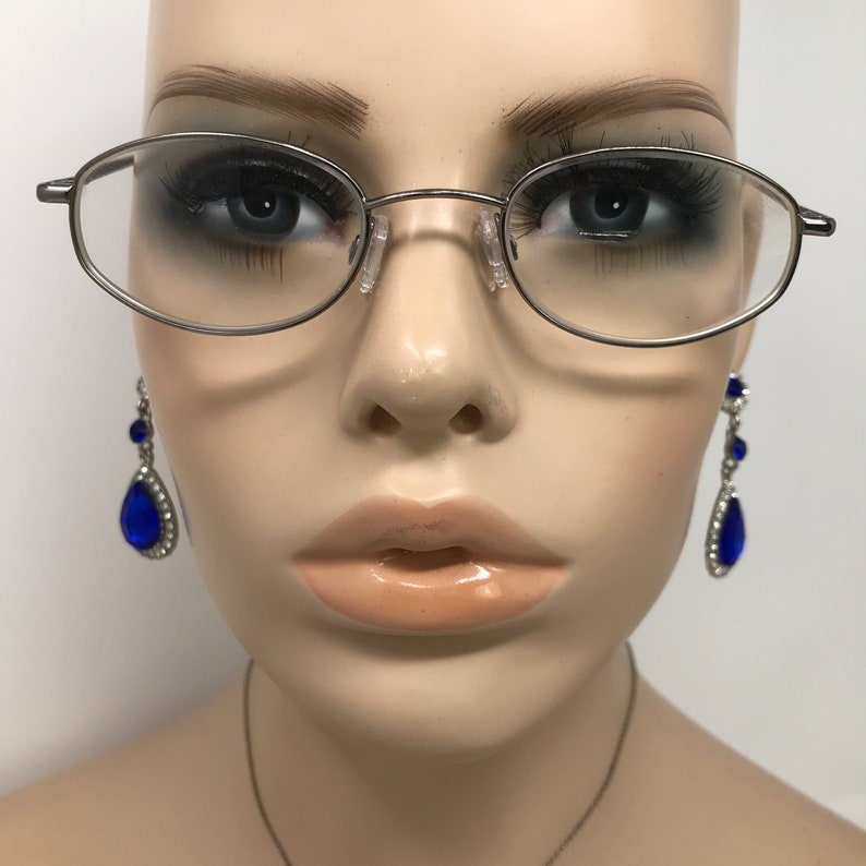 Op de afbeelding: Ovale bril met zilveren montuur en heldere glazen. De bril is op het gezicht van een mannequin geplaatst, gecombineerd met bungelende blauwe en zilveren oorbellen. De oorbellen hebben een druppelvormige blauwe edelsteen.