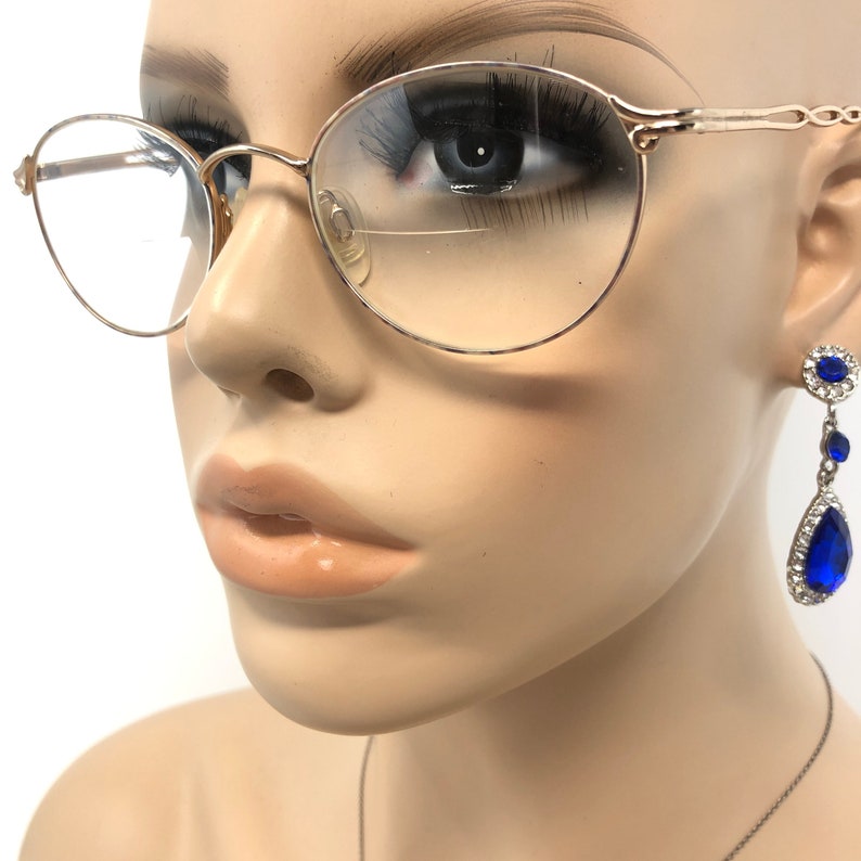 K&ouml;nnte beinhalten: Ein Schaufensterpuppenkopf mit goldgerahmter Brille und einem Paar baumelnder blauer und silberner Ohrringe. Die Brille hat einen d&uuml;nnen Rahmen und runde Gl&auml;ser. Die Ohrringe haben einen gro&szlig;en, tropfenf&ouml;rmigen blauen Stein.