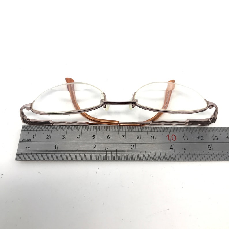 K&ouml;nnte beinhalten: Eine braune und silberne Brille mit einem Metallrahmen und klaren Gl&auml;sern. Die Brille wird mit einem Lineal gemessen und zeigt eine Breite von 10 Zentimetern.
