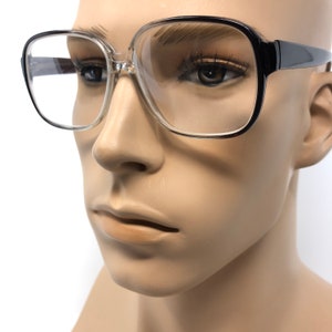 K&ouml;nnte beinhalten: Brille mit transparentem Rahmen, schwarzen B&uuml;geln und quadratischer Form. Die Brille befindet sich auf einem Schaufensterpuppenkopf. Die Gl&auml;ser sind klar und die Fassungen sind dick und k&uuml;hn, was einen Retro-Look ergibt.