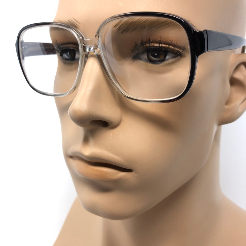 K&ouml;nnte beinhalten: Brille mit transparentem Rahmen, schwarzen B&uuml;geln und quadratischer Form. Die Brille befindet sich auf einem Schaufensterpuppenkopf. Die Gl&auml;ser sind klar und die Fassungen sind dick und k&uuml;hn, was einen Retro-Look ergibt.