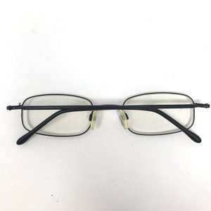 Puede incluir: Gafas rectangulares negras con lentes transparentes. Las gafas tienen una montura negra fina y peque&ntilde;as almohadillas nasales. Las patillas tambi&eacute;n son negras y se curvan en los extremos.