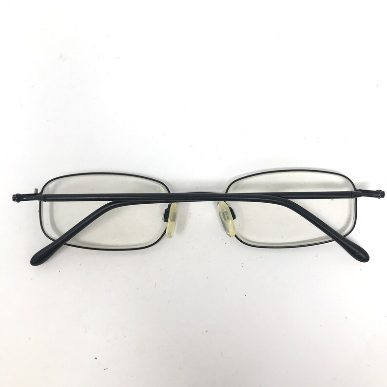 Puede incluir: Gafas rectangulares negras con lentes transparentes. Las gafas tienen una montura negra fina y peque&ntilde;as almohadillas nasales. Las patillas tambi&eacute;n son negras y se curvan en los extremos.