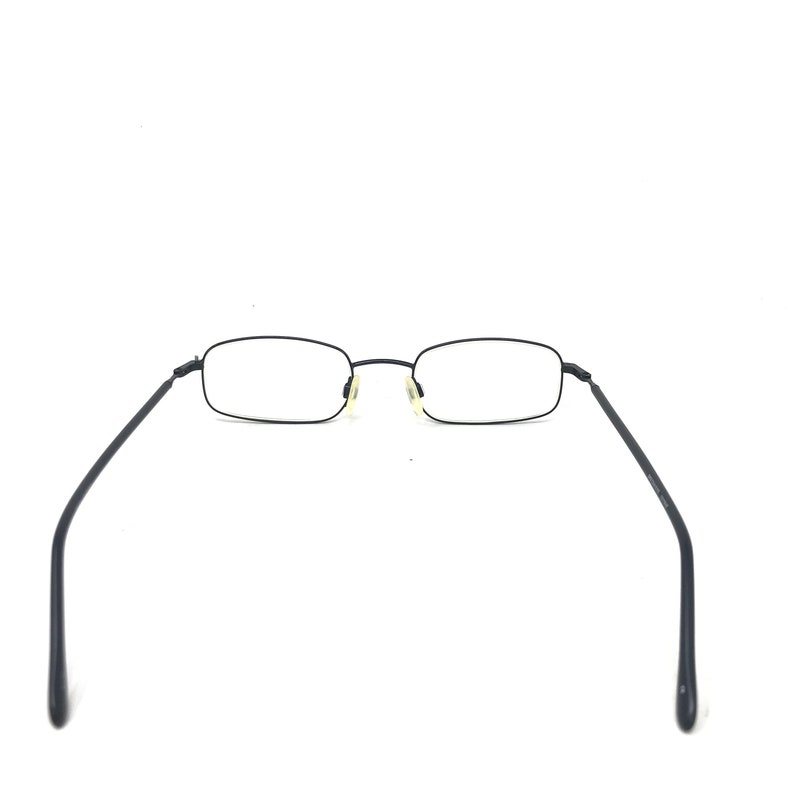 Puede incluir: Gafas rectangulares negras con lentes transparentes. Las gafas tienen una montura y patillas finas y oscuras que se extienden hacia afuera. Las almohadillas nasales son de color amarillo claro.