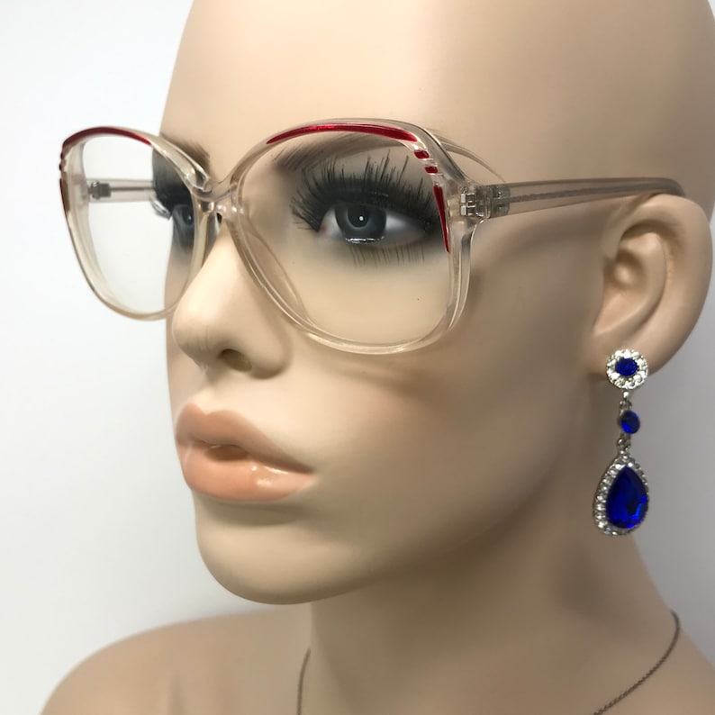 May include: Clear eyeglasses with a red accent on the top frame. The glasses are square-shaped and have a vintage style. A pair of blue and silver drop earrings are also visible.