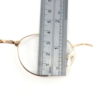 May include: Close-up of gold-rimmed eyeglasses with a ruler showing a measurement of approximately 1.5 inches. The glasses have a classic round shape and clear lenses. The ruler is marked in inches and centimeters.
