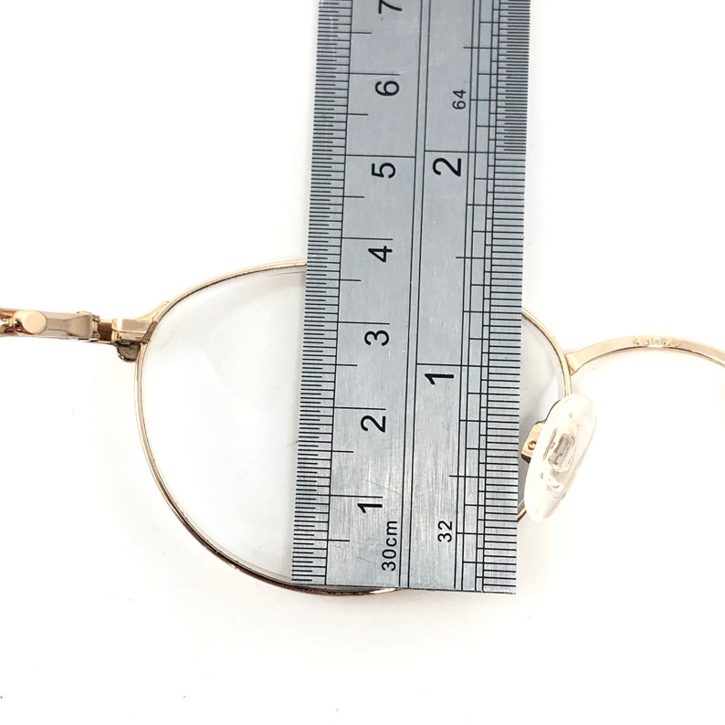 May include: Close-up of gold-rimmed eyeglasses with a ruler showing a measurement of approximately 1.5 inches. The glasses have a classic round shape and clear lenses. The ruler is marked in inches and centimeters.