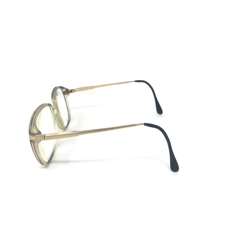 May include: A pair of eyeglasses with a light gray frame and clear lenses. The temples are gold-colored with black ear pieces. The glasses are set against a white background.