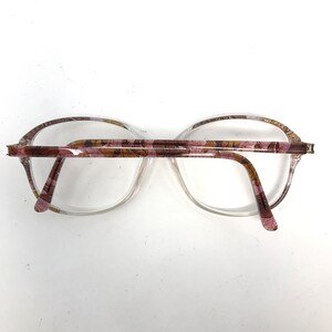 May include: Clear eyeglasses with a floral pattern on the temples and upper rims. The frames are a light pink and brown color, with a clear lens. The glasses are set against a white background.