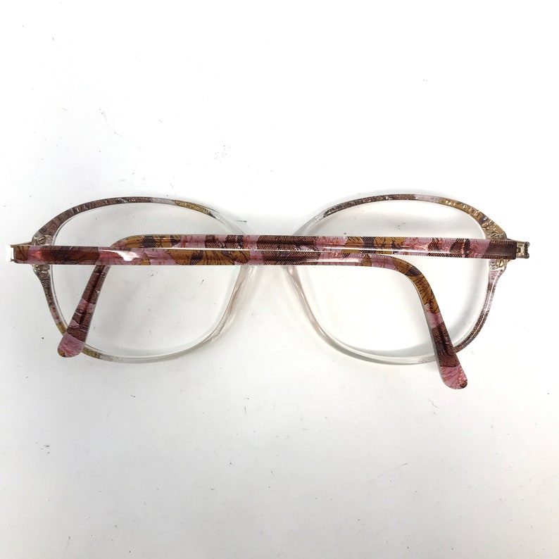 May include: Clear eyeglasses with a floral pattern on the temples and upper rims. The frames are a light pink and brown color, with a clear lens. The glasses are set against a white background.