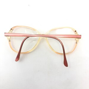 Puede incluir: Gafas vintage con montura y varillas de color rosa claro. Las lentes son transparentes y la montura presenta un dise&ntilde;o retro sutil. Las gafas se presentan sobre un fondo blanco, resaltando su forma y color.