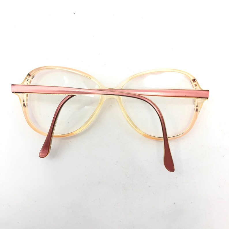 Puede incluir: Gafas vintage con montura y varillas de color rosa claro. Las lentes son transparentes y la montura presenta un dise&ntilde;o retro sutil. Las gafas se presentan sobre un fondo blanco, resaltando su forma y color.