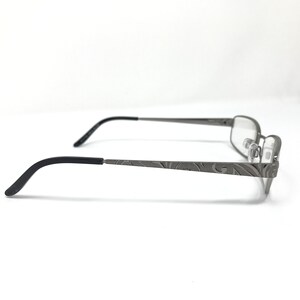 May include: Gray metal eyeglasses with a rectangular frame and black temples. The temples feature an etched floral design. The glasses are shown in profile against a white background.