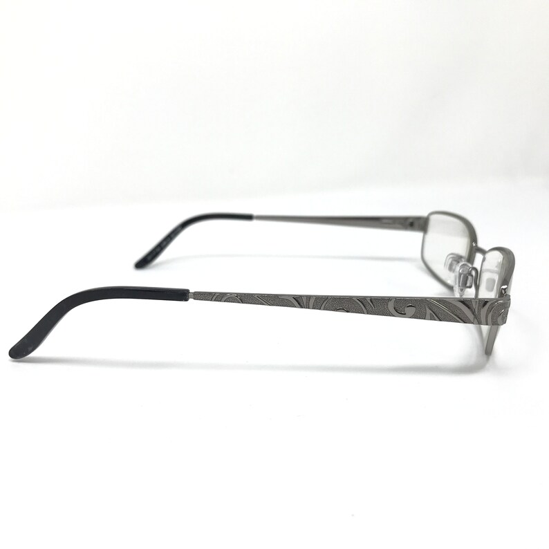 May include: Gray metal eyeglasses with a rectangular frame and black temples. The temples feature an etched floral design. The glasses are shown in profile against a white background.