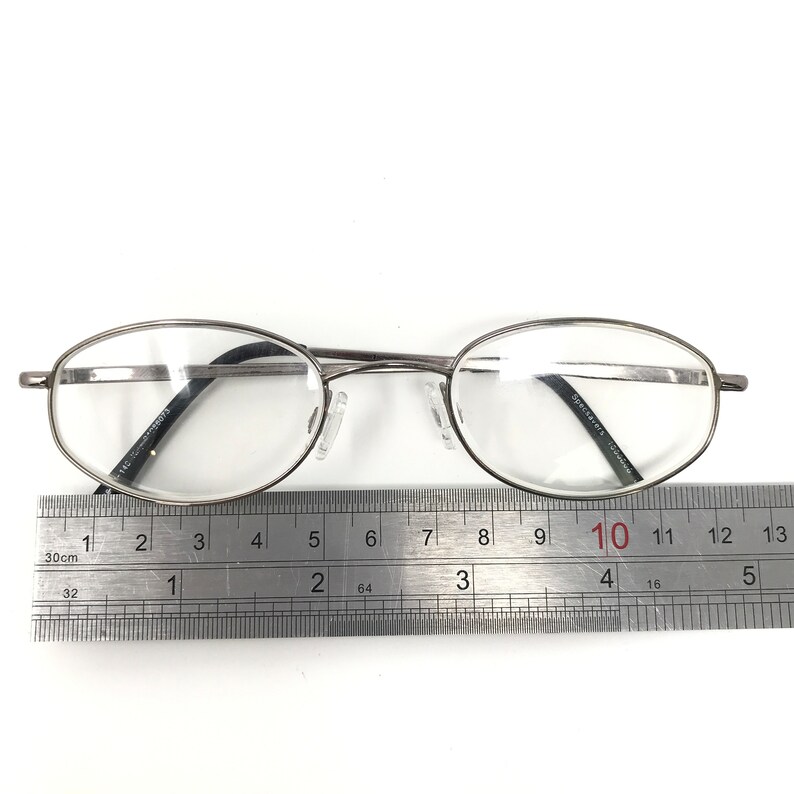 Op de afbeelding: Zilverkleurige bril met ovale glazen. De bril ligt op een liniaal, het montuur meet ongeveer 13 cm. De glazen zijn helder en de neuspads zijn wit.