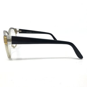 May include: Clear-framed eyeglasses with black temples. The glasses have a rectangular shape and a gold-colored accent on the frame. The text "SPECSAVERS" is printed on the temple.