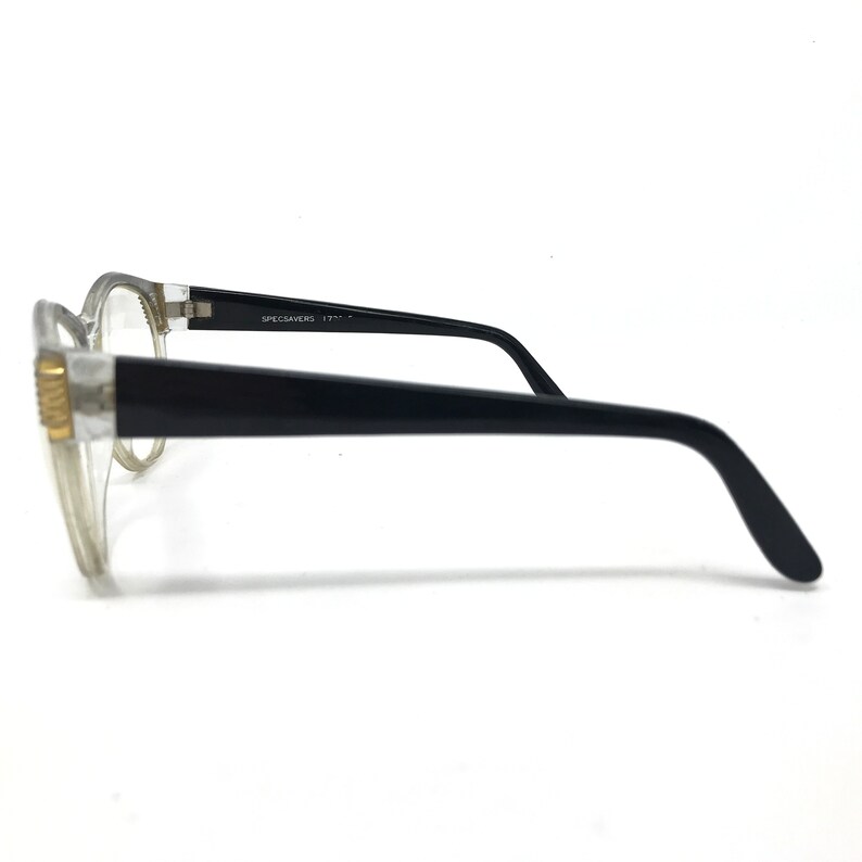 May include: Clear-framed eyeglasses with black temples. The glasses have a rectangular shape and a gold-colored accent on the frame. The text "SPECSAVERS" is printed on the temple.