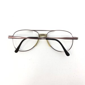 May include: Aviator-style eyeglasses with a brown metal frame and clear lenses. The glasses have a double bridge and black temples. The lenses are oval-shaped and the nose pads are a light beige color.