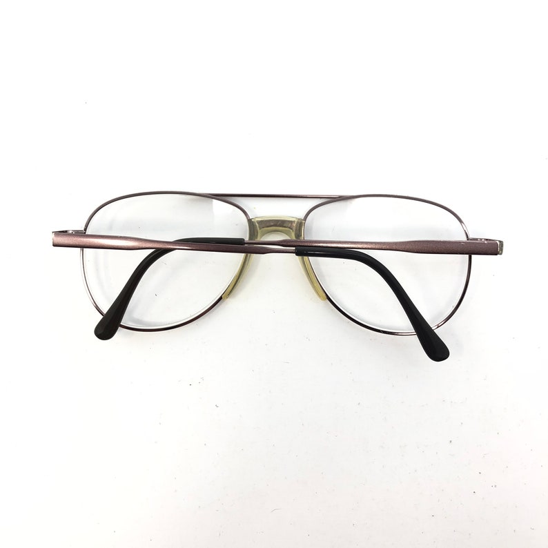 May include: Aviator-style eyeglasses with a brown metal frame and clear lenses. The glasses have a double bridge and black temples. The lenses are oval-shaped and the nose pads are a light beige color.