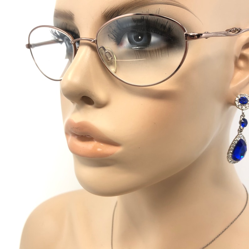 May include: Rose gold oval eyeglasses with clear lenses and a delicate frame. The mannequin also wears a pair of dangling earrings with blue teardrop-shaped gems and clear crystals. The earrings are set in silver-colored metal.