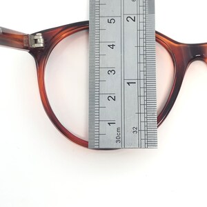 May include: Close-up of a pair of brown tortoiseshell eyeglasses with a ruler measuring the frame's height. The frame is approximately 2 inches (5 cm) tall. The glasses are set against a white background.