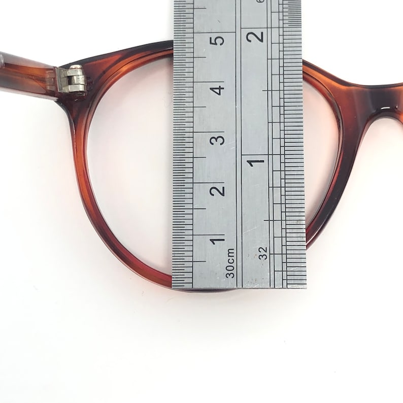 May include: Close-up of a pair of brown tortoiseshell eyeglasses with a ruler measuring the frame's height. The frame is approximately 2 inches (5 cm) tall. The glasses are set against a white background.