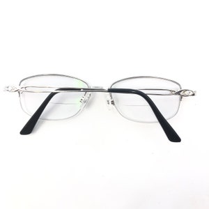 May include: Silver-framed reading glasses with black temples and clear lenses. The glasses have a half-rim design and decorative accents on the temples. The lenses have a bifocal design.