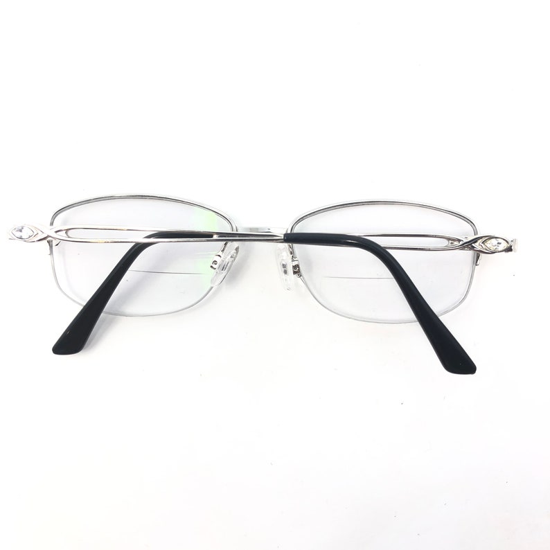 May include: Silver-framed reading glasses with black temples and clear lenses. The glasses have a half-rim design and decorative accents on the temples. The lenses have a bifocal design.
