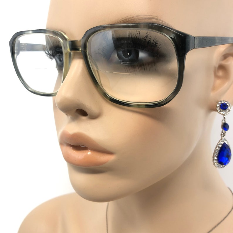 May include: Close-up of eyeglasses with a gray and green speckled frame. The glasses have a square shape and clear lenses. A blue and silver drop earring is visible on the right side of the image.