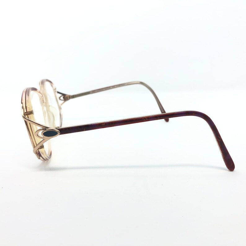 K&ouml;nnte beinhalten: Vintage Brille mit hellbraunem Rahmen und klaren Gl&auml;sern. Die B&uuml;gel sind dunkelbraun mit einem dezenten Muster. Die Fassungen haben eine einzigartige Form mit einem dunkelblauen ovalen Akzent an der Seite.