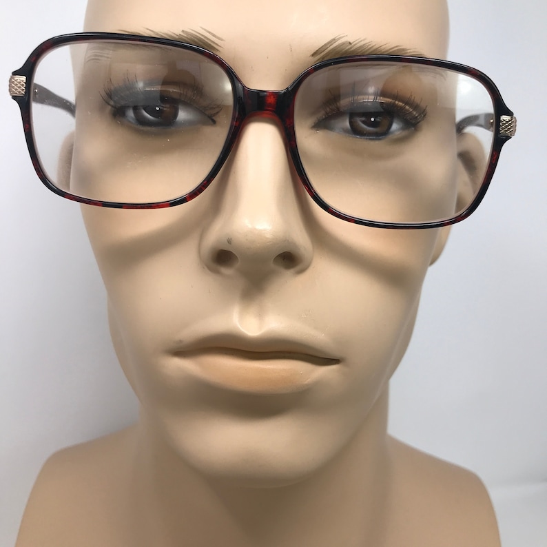 May include: Square-framed eyeglasses with a tortoiseshell pattern on the rims. The glasses have clear lenses and gold-colored accents on the temples. The frames are set against a neutral background.