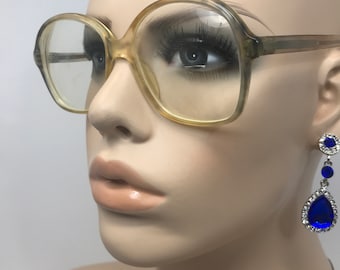 May include: A mannequin wearing oversized, light-colored eyeglasses with a rounded frame. The mannequin also wears dangling earrings with blue teardrop-shaped gems and smaller blue gems. The background is plain white.