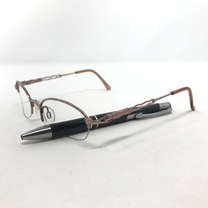 May include: A pair of eyeglasses with a brown metal frame and a black pen. The eyeglasses have a delicate, wire-like design on the frame. The pen is black with a silver tip and clip. The items are on a white surface.