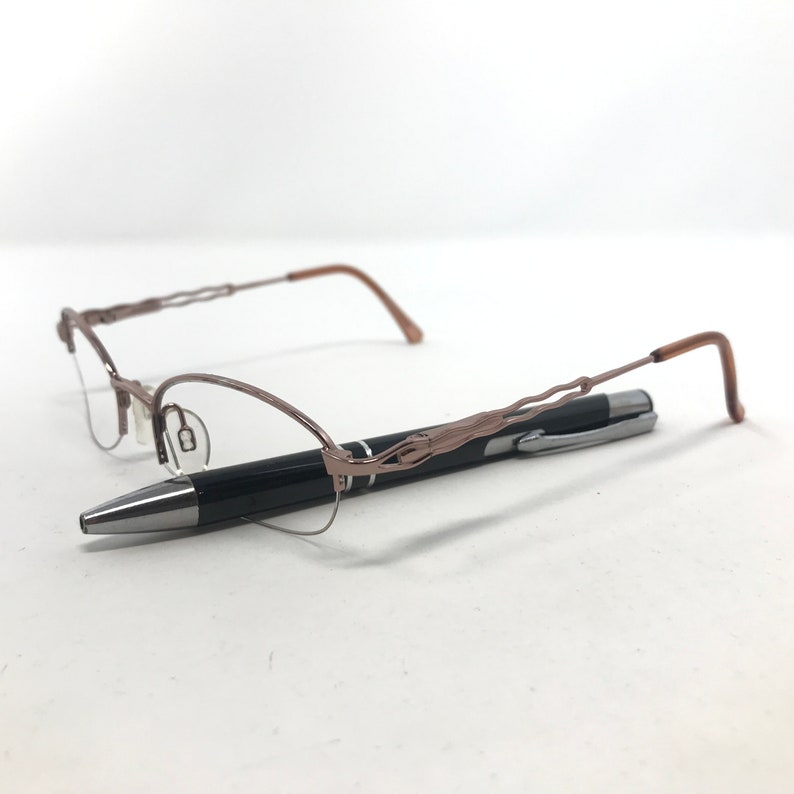 May include: A pair of eyeglasses with a brown metal frame and a black pen. The eyeglasses have a delicate, wire-like design on the frame. The pen is black with a silver tip and clip. The items are on a white surface.