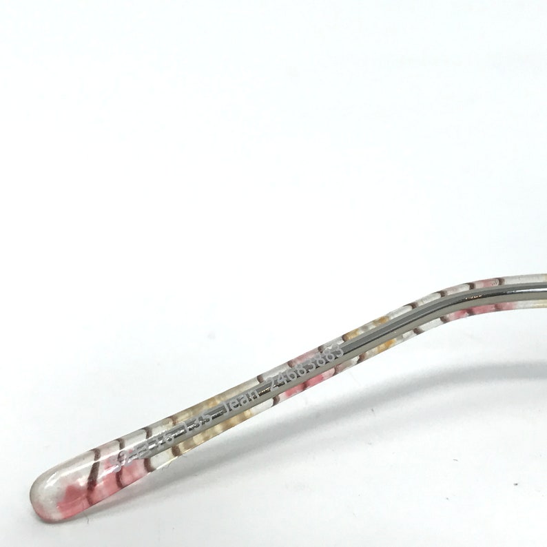 May include: Close-up of a glasses arm with a floral pattern in pink, white, and yellow. The arm is clear with a silver metal frame. The text "jean 24665869" is printed on the arm.