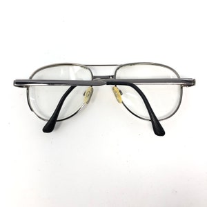 Peut inclure: Lunettes de vue aviateur &agrave; monture argent&eacute;e avec verres transparents. Les lunettes ont un double pont et des branches noires. Les plaquettes de nez sont de couleur jaune clair. Sur fond blanc.