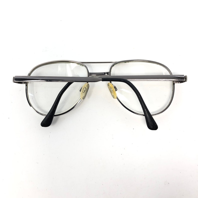 Peut inclure: Lunettes de vue aviateur &agrave; monture argent&eacute;e avec verres transparents. Les lunettes ont un double pont et des branches noires. Les plaquettes de nez sont de couleur jaune clair. Sur fond blanc.