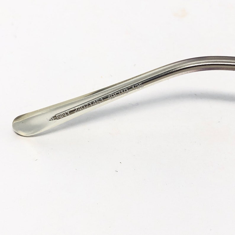 May include: Close-up of the temple arm of eyeglasses. The arm is silver-toned metal with a clear, rounded tip. The text "JENNY 2407 145 49[]19 135" is printed on the arm.
