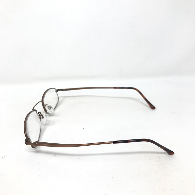 May include: A pair of eyeglasses with a brown metal frame and clear lenses. The glasses have a classic, minimalist design with a rectangular shape. The temples have a tortoiseshell pattern at the ends.
