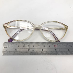 May include: Clear oval eyeglasses with a gold-colored metal frame and purple accents. The glasses are placed on a white surface next to a ruler. The glasses measure approximately 5 inches in length.