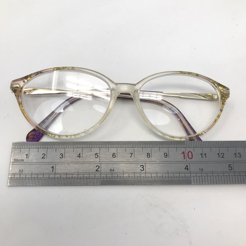 May include: Clear oval eyeglasses with a gold-colored metal frame and purple accents. The glasses are placed on a white surface next to a ruler. The glasses measure approximately 5 inches in length.