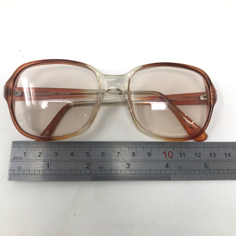 May include: A pair of eyeglasses with a clear and brown gradient frame. The glasses have a rectangular shape and are placed on a white surface next to a ruler. The text "SPECSAVERS 1754205" is visible on the frame.