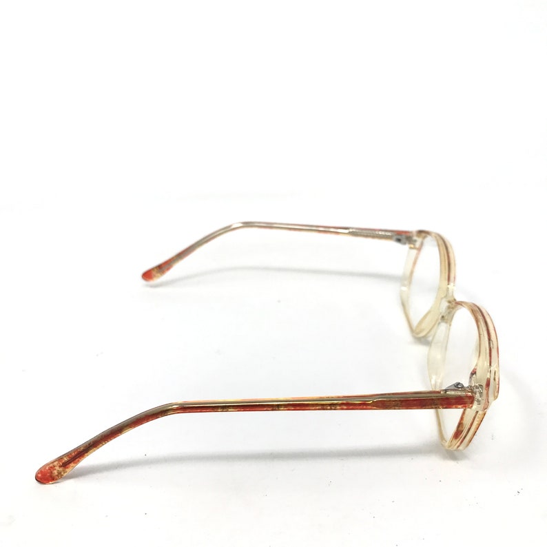 May include: A pair of vintage eyeglasses with a clear, rectangular frame and thin, gold-colored metal arms. The arms have a reddish-brown pattern. The glasses are set against a white background.