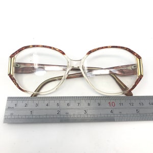 May include: Clear eyeglasses with a geometric frame, featuring brown and gold accents on the upper rims and temples. The glasses have a rectangular shape with a ruler placed below, measuring the frame's width.