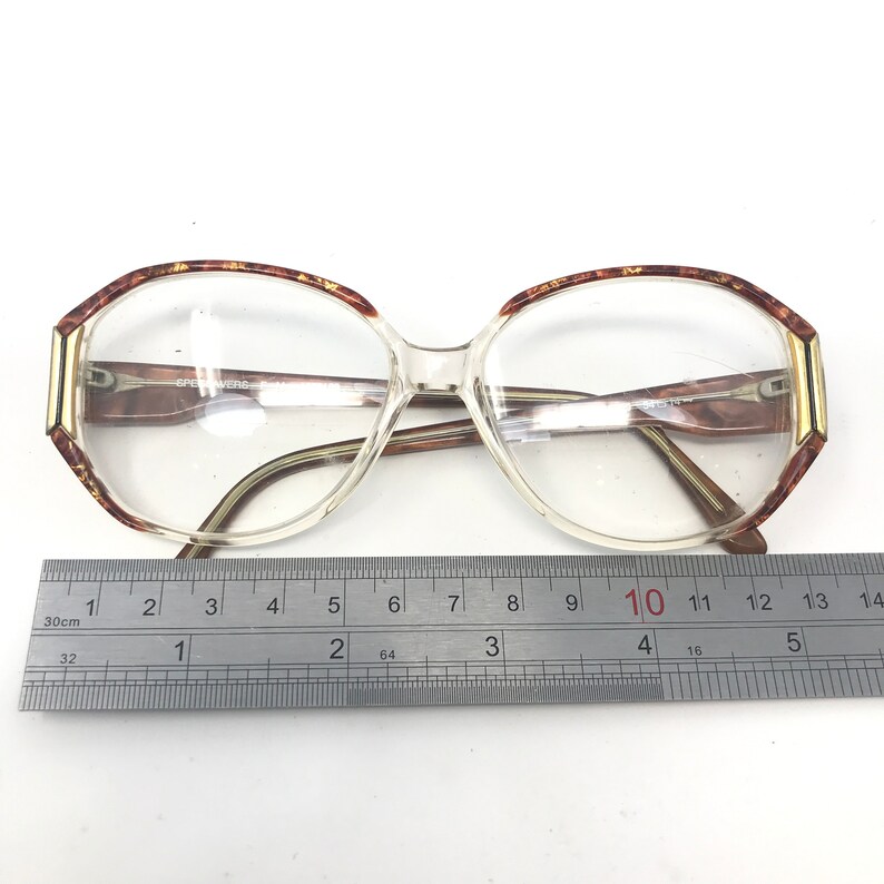 May include: Clear eyeglasses with a geometric frame, featuring brown and gold accents on the upper rims and temples. The glasses have a rectangular shape with a ruler placed below, measuring the frame's width.