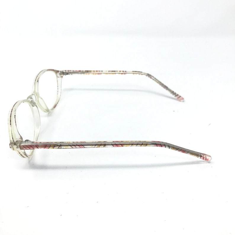 May include: Clear eyeglasses with round lenses and patterned temples. The temples feature a repeating pattern of red, yellow, and brown lines. The glasses are set against a white background.