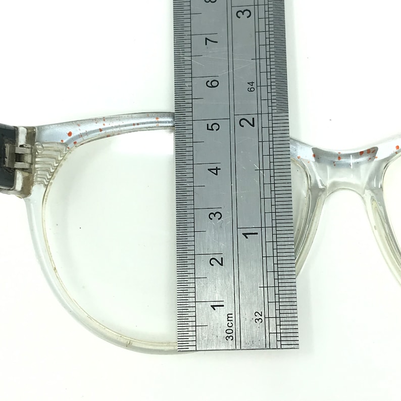 May include: Clear eyeglasses with a ruler measuring the frame. The glasses have a transparent frame with small red flecks. The ruler shows a measurement of approximately 5 inches.