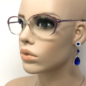 May include: A pair of clear, rectangular eyeglasses with a light purple frame. The eyeglasses are on a mannequin's face. The mannequin is wearing blue teardrop-shaped earrings with rhinestones.