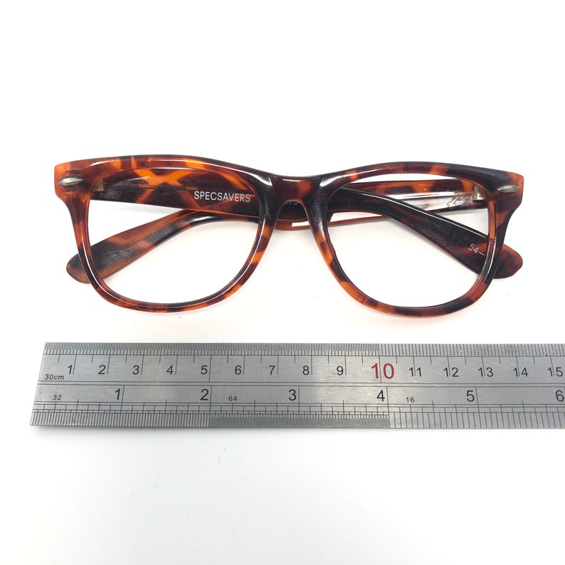 K&ouml;nnte beinhalten: Brille mit Schildpattmuster und dem Text "SPECSAVERS" auf dem Rahmen. Die Brille liegt neben einem Lineal, mit einer Rahmenbreite von etwa 14 cm.